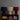Three pleated fabric table lamps in blue, beige, and red on a wooden shelf with books against a gray wall.