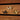 Three decorative hooks shaped like stylized letters or abstract shapes, shown on a wooden drawer front. The pulls are finished in silver, black with gold trim, and gold.