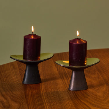 Two purple candles on black holders on a wooden table with a green wall background
