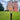A tall, dark metal bollard post with a tiered top stands on a green grass lawn in front of a large red brick castle under a cloudy blue sky.