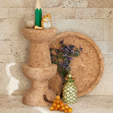 Decorative items including a pineapple, small animal figurine, and fruit on a stone pedestal against a textured wall.