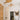 An art gallery or studio interior with a large beige tapestry on the left wall, a vertical cream text banner, butterfly artwork, hanging wooden butterfly forms, studio lights, and a table covered with printed materials in the foreground.