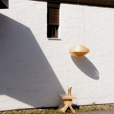 Modern pendant light hanging against a white wall with a small wooden stool. Hans-Agne Jakobsson '1003 Hans Pendant' in Pine for Two Enlighten