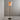 A modern floor lamp with a tapered beige fabric shade, a slim gold metal pole, a black central accent, and a round black base, shown lit in a minimalist gray room corner.