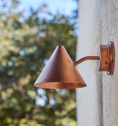 Copper outdoor wall light fixture on a concrete wall with blurred greenery in the background