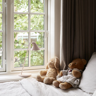 Two teddy bears on a bed next to a large window with curtains and a pale pink lamp