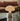 Mushroom-shaped lamp on a glass table with a brown sofa and plant in the background
