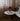 Round white table with a red lamp and books in a room with curtains and a sofa.
