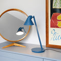 Blue desk lamp on a surface with a mirror and artwork in the background