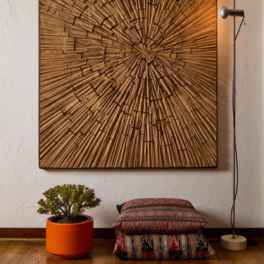 Wooden wall art with concentric circle design on a white wall, next to a plant in an orange midcentury gainey planter and a cushion.