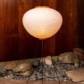 Round akari 10A wash paper japanese floor lamp with a warm glow on a tripod stand against a wooden wall.
