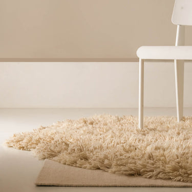 Beige  deep shag rug on a light wooden floor with a white chair against a beige wall.