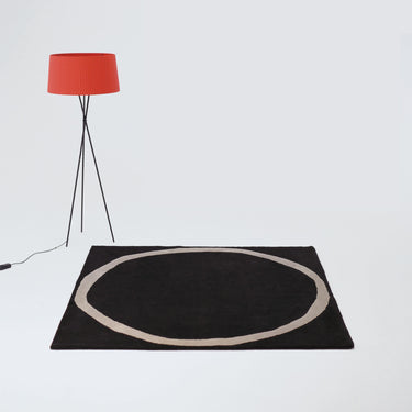 Black rug with a circular design on a white floor, featuring a red lamp.