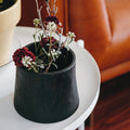 Black brass vase or planter with dried flowers on a white table in a living room setting.