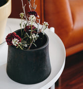 Black brass vase or planter with dried flowers on a white table in a living room setting.