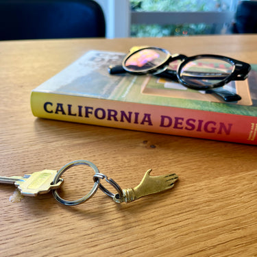 Keychain with a hand design on a wooden table with a book titled 'California Design' in the background.