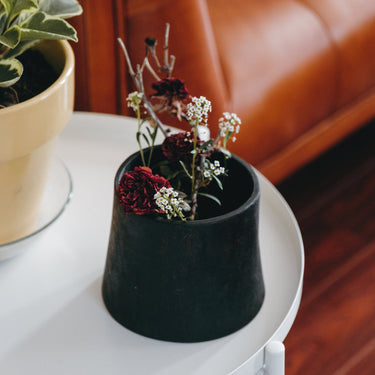 Black planter with flowers on a white table next to a brown leather couch.