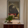 Framed poster titled 'EXACTITUDE ETAT' on a wall above a wooden cabinet with a lamp and plant.