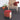 Color-blocked ottoman in front of a woven chair with a red blanket and potted plants on a gray carpet.