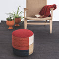 Color-blocked ottoman in front of a woven chair with a red blanket and potted plants on a gray carpet.