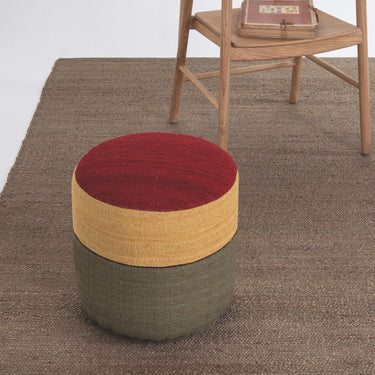 Multicolored ottoman on a brown carpet with a wooden chair in the background.