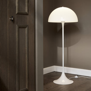White 70s floor lamp against a dark wall with wooden floor and door.
