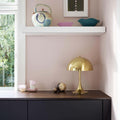 Decorative setup with a gold 1970s brass panton lamp, ceramic bowls, and a framed picture on a white shelf against a pink wall.