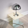 Modern silver table lamp on a glass cube with a chair in the foreground.