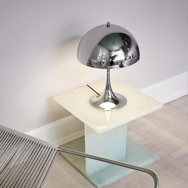 Modern silver table lamp on a glass cube with a chair in the foreground.