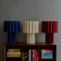 Three pleated fabric table lamps in blue, beige, and red on a wooden shelf with books against a gray wall.