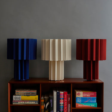 Three pleated fabric table lamps in blue, beige, and red on a wooden shelf with books against a gray wall.