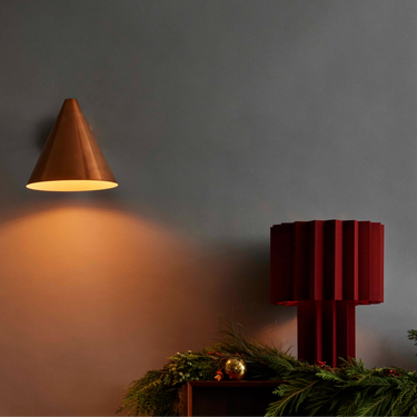 Conical wall light fixture casting a warm glow on a dark gray wall with a red table lamp and greenery.