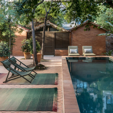 Outdoor setting with lounge chairs and rugs by a pool, surrounded by trees and buildings.