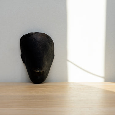 Black mask sculpture on a wooden surface with a light gray wall in the background