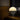 A modern table lamp with a glowing white dome shade and a silver wire spiral base, shown lit on a wooden shelf in a dimly lit interior setting.