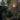 Person watering plants in a garden with a brick wall and outdoor light fixture.
