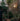 Person watering plants in a garden with a brick wall and outdoor light fixture.